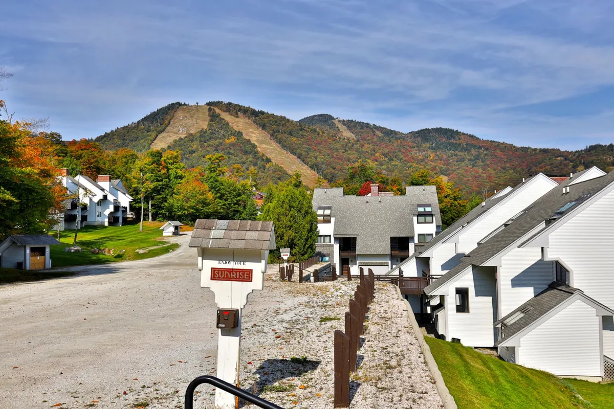 This exterior image showcases residential buildings nestled against the backdrop of Killington's impressive mountain vista, with a road leading up to the area. The scene conveys the proximity to the slopes and the natural beauty of the setting. Viewers can imagine stepping out their door and heading straight onto the ski trails or hiking paths.