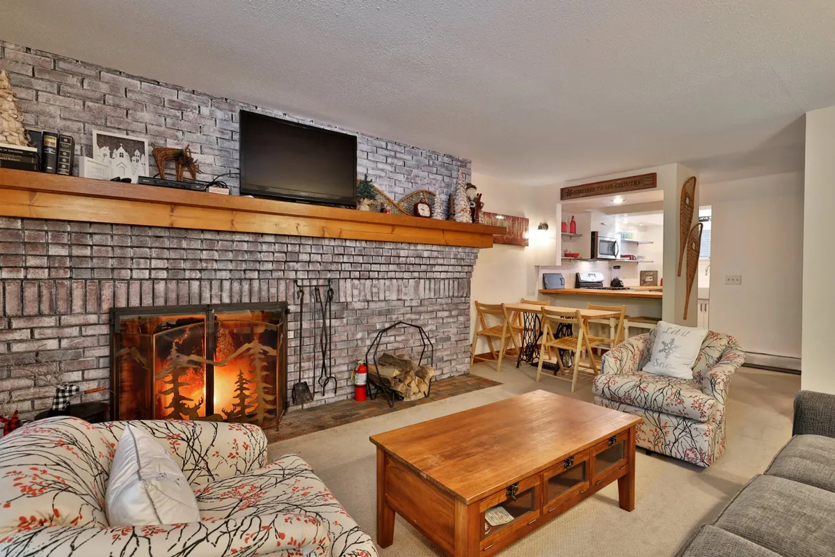 The image shows a warm and inviting living space with a stone fireplace as the centerpiece, a rustic wooden mantel, a comfortable seating area, and a dining table ready for shared meals. The ambiance suggests relaxation, warmth, and togetherness after a day outdoors.