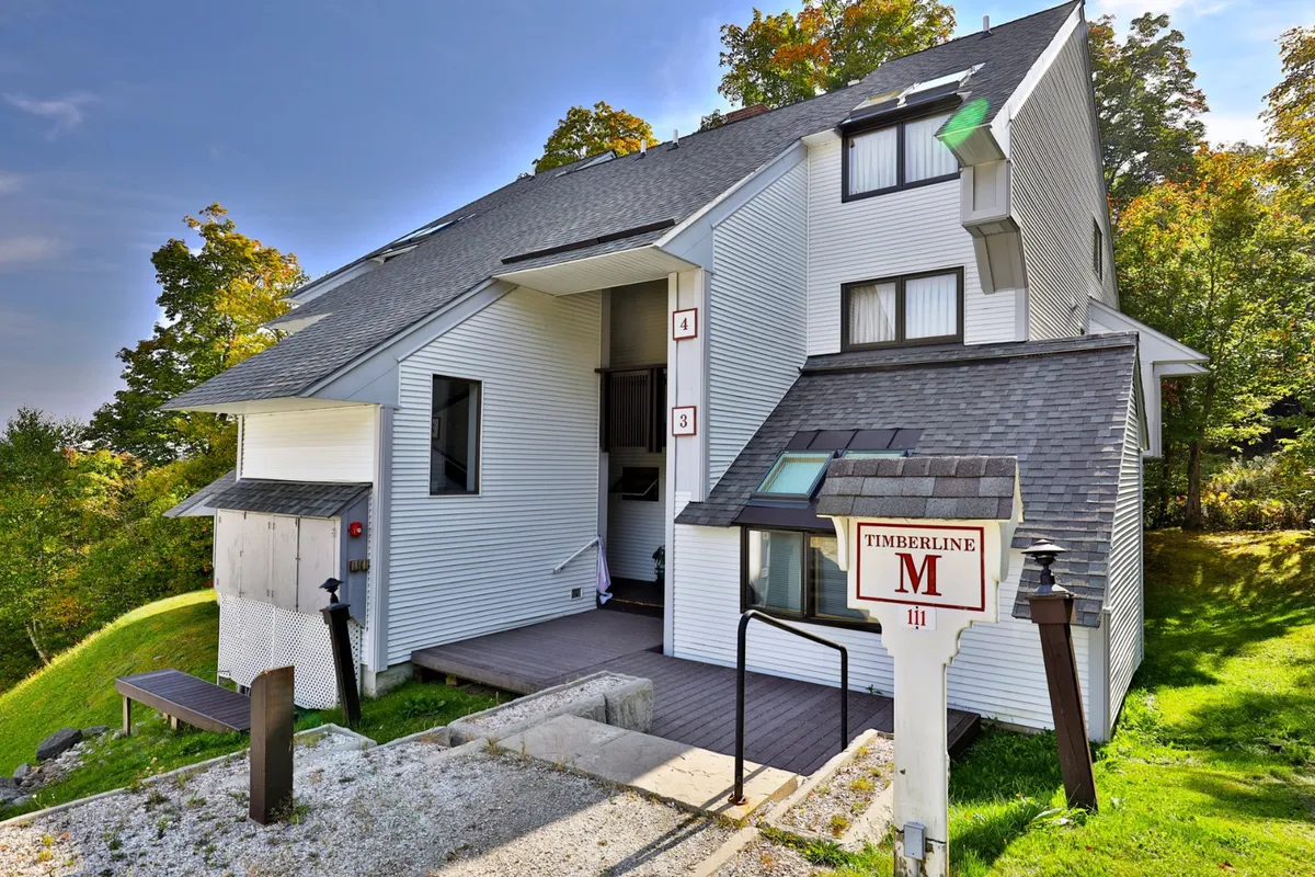 An inviting exterior shot of the property entrance, featuring the Timberline sign, surrounding greenery, and a welcoming porch area. The scene suggests a friendly, communal atmosphere and sets the stage for returning home after a day of adventure.