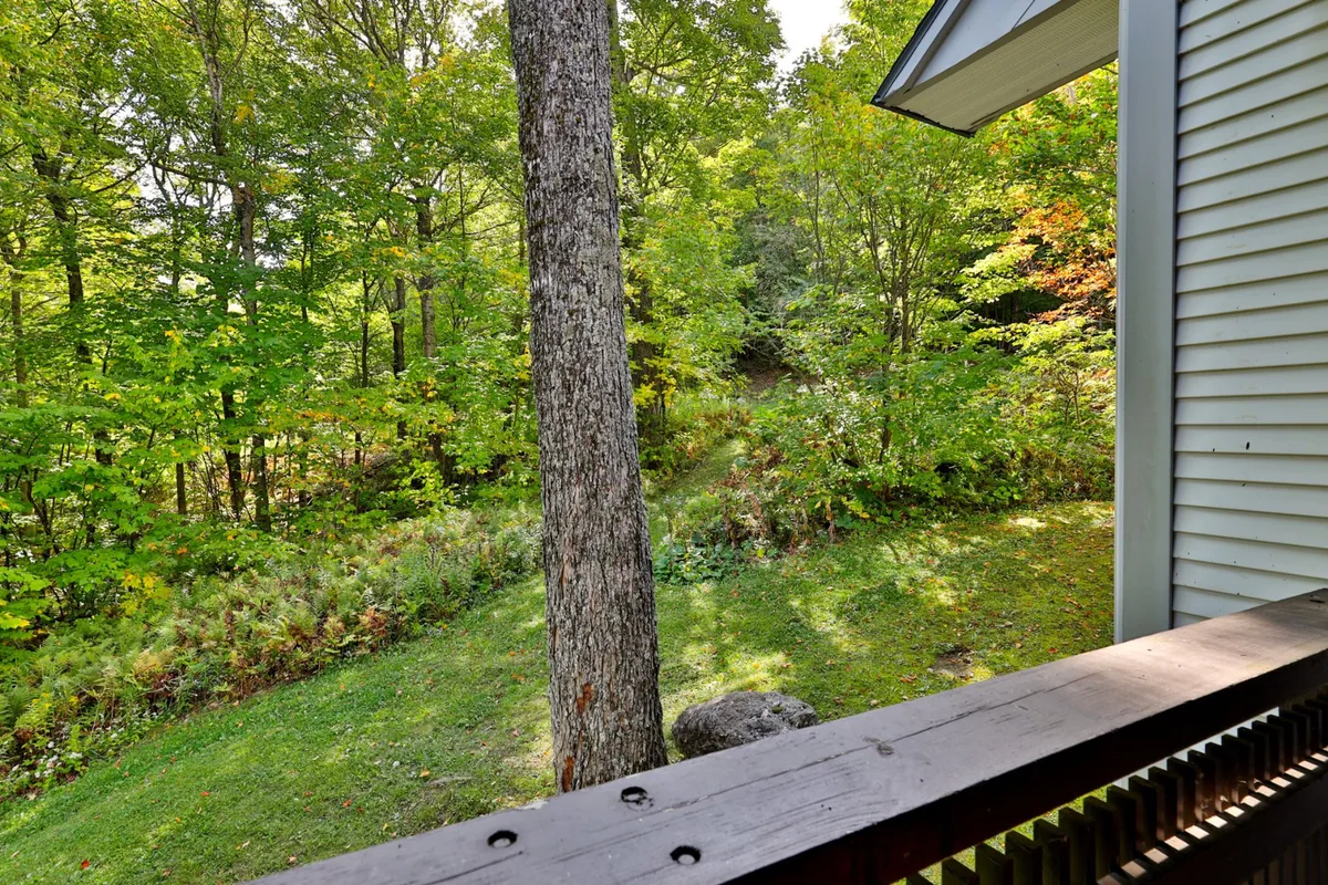 This exterior image features lush greenery, tall trees, a wooden railing, stone, and the side of a house nestled in nature. The tranquil setting suggests a strong connection to the outdoors and the serenity of the mountain environment.