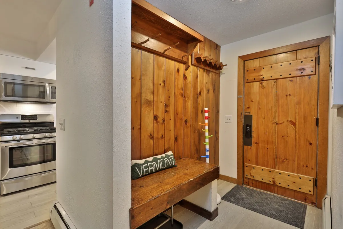 This image shows the entryway of the condo, featuring a wooden bench, the front door equipped with a smart lock, and a glimpse of the kitchen area. The setting is welcoming, practical, and highlights an organized arrival experience.