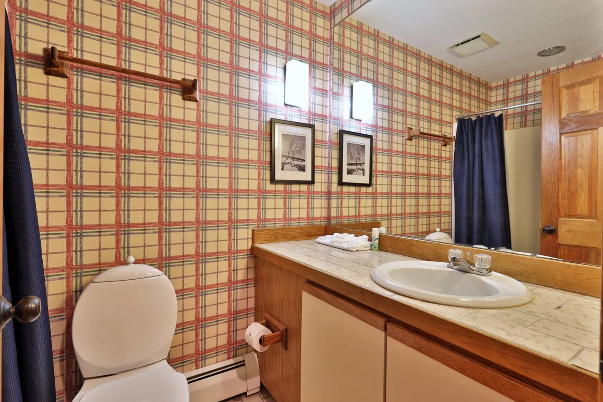 A well-lit bathroom with plaid wallpaper, a sink and countertop, framed photographs, and curtains. The inviting space is designed for personal grooming and getting ready for the day's activities.