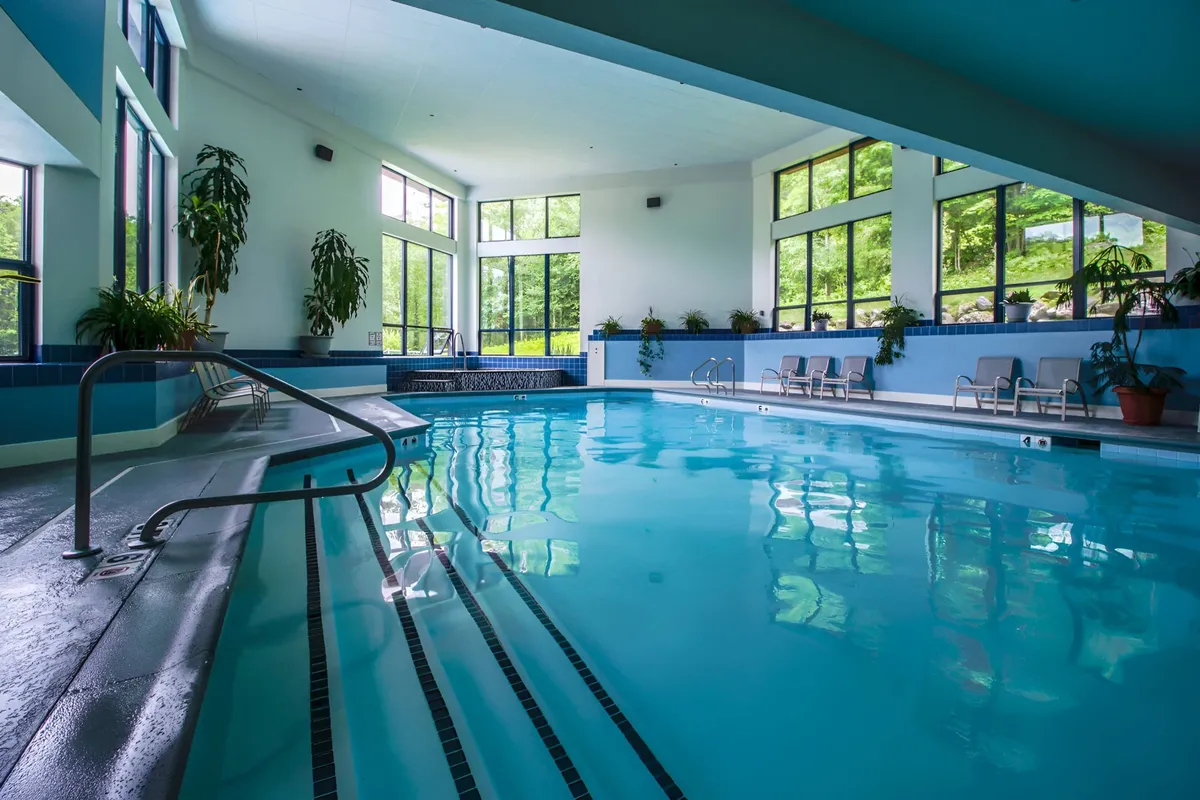 A serene indoor swimming pool area with large windows bringing in natural light, lush greenery accenting the exterior views, and lounge chairs lining the poolside. The setting is tranquil and inviting, perfect for swimming or relaxing by the water.