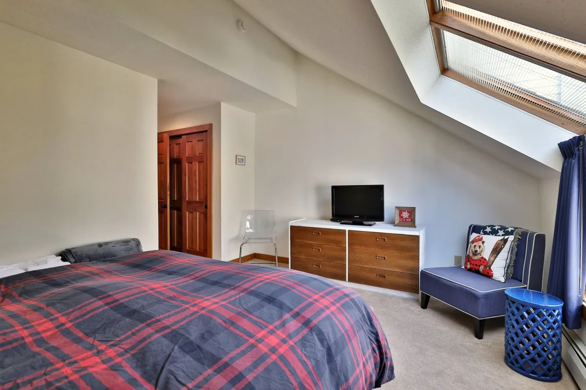 A cozy bedroom with a bed featuring plaid bedding, a wooden dresser, a TV, and a blue accent chair. The room is brightened by natural light, creating a comfortable and inviting space perfect for unwinding after a day of adventure.
